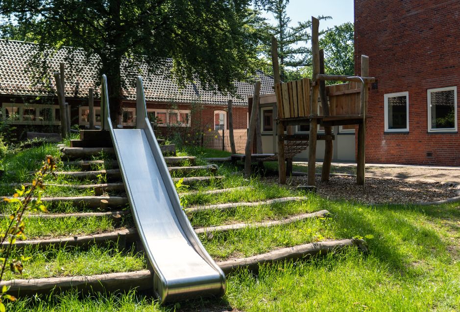 Spielplatz mit Rutsche und Holzklettergerüst vor einem Backsteingebäude.